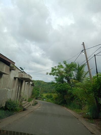 The image shows a street in Blangrheuem, Indonesia on an overcast day. The road is asphalt, curving slightly to the left and appearing damp. On the left side of the road is a partially visible, single-story, concrete building with a satellite dish on the roof. The building has a series of protruding concrete overhangs supported by columns. A short set of concrete steps lead up to the building. Lush green foliage grows along the roadside. On the right side of the road is a mix of trees and bushes. A concrete utility pole stands upright among the greenery, with wires extending from it. Further down the road, a glimpse of more green hills and vegetation is visible. The sky is filled with dense, grey clouds, indicating possible rain.