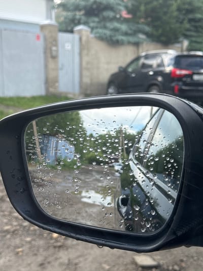 The image is a close-up of a car's side-view mirror, which is covered in raindrops. The mirror reflects a blurry scene of a street with puddles, trees, and buildings. In the background, there are two dark-colored SUVs parked on the side of the street. The overall impression is that of a rainy day in a residential area. The location is identified as Кусимовского рудника, Russia.