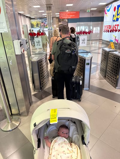 A top-down perspective shows a man in a black backpack, black pants, and grey t-shirt next to luggage with a child in a stroller with a Ryanair bag tag attached in an airport in the Italian city of Cinisi. The man is standing in front of the luggage carousel. The baby is awake and looking at the camera. Overhead signs in the airport indicate the baggage claim and elevator.