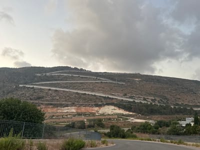 The image captures a wide, scenic view of a mountainous landscape on the outskirts of Shlomi, Israel. The terrain is arid and rocky, with sparse vegetation. A prominent feature is a winding road that snakes its way up the hillside, marked by a series of zig-zagging turns. Along the side of the road, a security fence runs intermittently, indicating the boundary of a secured area.
In the foreground, a dark metal fence with vertical bars runs horizontally across the bottom of the frame, separating the viewer from a grassy verge and some low-lying shrubs. Beyond this, a paved road curves into the scene from the right.
The hillside is characterized by a mix of dry scrub, scattered trees, and exposed rock faces, some of which show distinct layers of reddish-brown earth. Higher up the slope, more trees are visible, interspersed with patches of barren land. The very top of the hill is lined with what appears to be a continuous structure, possibly a wall or a series of fortifications, stretching across the ridge. A slender communication tower or antenna can be seen on the distant hilltop.
The sky is overcast with thick, grey clouds, suggesting a diffused light or possibly an impending change in weather. The overall impression is one of a rugged, natural environment with man-made infrastructure adapted to the challenging topography. The presence of the security fence strongly suggests this is a border region or an area with specific security considerations.