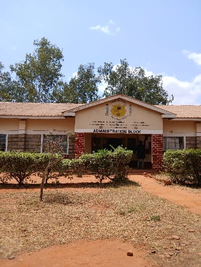 An exterior, daytime photograph depicts the administration block of an educational institution in Masii ward, Kenya. The single-story building features a light beige facade with a reddish-brown corrugated roof. The central section is distinguished by a gable inscribed with "ADMINISTRATION BLOCK" in black capital letters. Above this, a crest displays a yellow circular emblem, possibly including "MASII" and "PRIMARY," flanked by two stylized figures: a male figure with a shield and spear on the left, and a female figure on the right. The lower portion of the building, particularly around the recessed entrance and beneath windows, is clad in red brickwork. Green, manicured shrubs line the base of the building. One individual, wearing dark clothing, is partially visible from behind, standing within the main doorway of the administration block. The foreground consists of reddish-brown earth with sparse, dry grass and a narrow dirt path leading to the entrance. A small, thin tree stands in the foreground, left of the path. Behind the building, several large trees with green foliage are visible against a light blue sky with scattered white clouds.