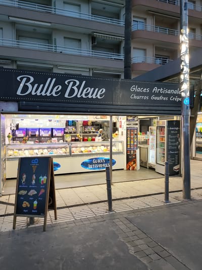 A storefront with a prominent sign reading "Bulle Bleue" is visible. The sign also advertises "Glaces Artisanales, Churros Gaufres Crépe". The storefront appears to be an ice cream shop or dessert parlor, with display cases filled with various ice creams. A menu board with illustrations of desserts and drinks, including ice cream cones, cups, crepes, and bubble tea, is placed in front of the shop. The shop is situated on a paved walkway, with metal bollards lining the path. In the background, a multi-story building with balconies is visible. The overall scene suggests an outdoor commercial area, likely in a tourist or pedestrian-friendly location. Based on the advertising in French, the location is identified as Canet-en-Roussillon, France.
