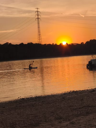 A lone figure kayaks on a calm body of water during sunset. The sun, a bright orange orb, is partially obscured by a treeline in the distance, casting a warm glow across the scene and reflecting on the water's surface. A tall electrical pylon stands prominently against the dusky sky, with power lines stretching across the frame. The shoreline in the foreground is composed of sand and small stones. A portion of a boat is visible on the right side of the image. This scene is likely captured at or near the Dunakeszi shores of the Danube River in Dunakeszi, Hungary.