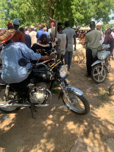 The image depicts an outdoor scene, likely a market, with a group of people and livestock. In the foreground, a motorcycle is parked, with its engine and front wheel clearly visible. A person wearing a blue shirt is seated on the motorcycle. Behind the motorcycle, several individuals are standing, some wearing traditional headwear and clothing. Sheep and goats are scattered among the people, suggesting a livestock market. The environment appears to be dusty with trees providing shade. The background shows more people and structures, hinting at a bustling gathering. The location is identified as Boi A, Nigeria.