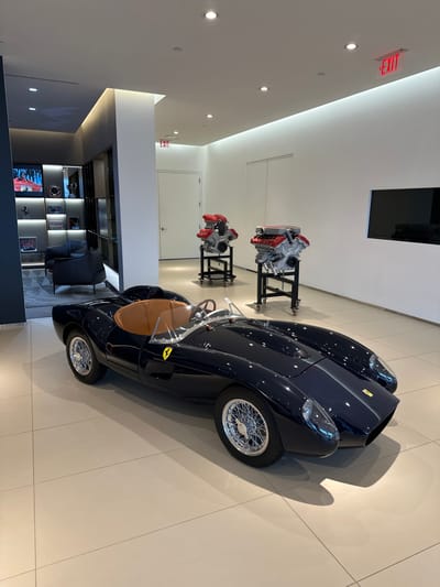 This image depicts a pristine, brightly lit showroom or exhibition space in Greenwich, United States, dedicated to automotive display. The central subject is a dark blue, vintage-style convertible sports car, resting on a light-colored tiled floor. The car exhibits classic features including a wooden steering wheel, a tan interior, and elegant chrome wire-spoke wheels. A distinctive yellow Ferrari shield emblem is visible on the side of the vehicle, indicating its heritage or inspiration.
In the background, two impressive automotive engines are showcased on black stands. These engines are characterized by their prominent red-colored tops, clearly displaying the "Ferrari" name, and highlight the brand's engineering. The showroom itself boasts clean white walls and ceiling, illuminated by multiple recessed spotlights and linear strip lighting, contributing to a modern and sophisticated atmosphere. A red "EXIT" sign is affixed to the ceiling in the upper left of the frame.
Towards the left and further into the room, a contemporary lounge area can be seen, furnished with a dark armchair, a low table, and a stylish shelving unit. This unit displays an assortment of automotive memorabilia, including trophies, scale models, and framed artwork. A small television screen within the shelving unit appears to show racing footage. The entire scene suggests a high-end environment for the appreciation of luxury automobiles, likely a Ferrari dealership, museum, or an exclusive private collection. No individuals are present in the image.
