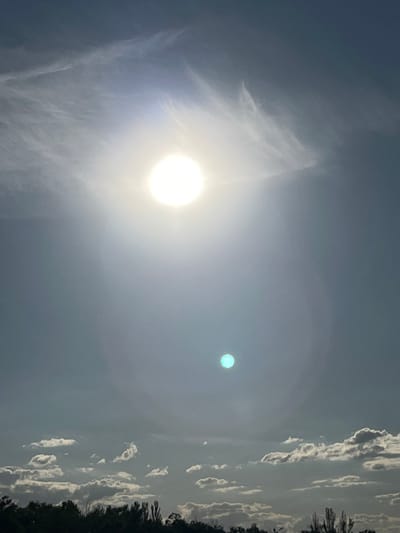 The image captures a bright sun in a partly cloudy sky, casting a lens flare that creates a secondary, smaller, aqua-colored orb below it. The scene is set against a backdrop of wispy clouds at higher altitudes and more defined cumulus clouds closer to the horizon. The bottom of the image shows a dark silhouette of trees, suggesting a natural or semi-rural environment. The overall lighting indicates it is daytime, with the sun being the primary light source. This scene is located in the vicinity of Shyroka Dacha, Ukraine.