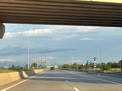 The image captures a highway view from under an overpass in Urbnisi, Georgia. The scene is dominated by a multi-lane highway stretching into the distance under a partly cloudy sky. The road surface is asphalt with clear white lane markings. A concrete barrier runs along the right side of the road.
Several vehicles are visible on the highway. In the foreground, there are no people. Further down the road, a white truck and a dark-colored car are traveling in the same direction. To the right, there are road signs, including a directional arrow sign and a sign with chevron markings.
On the left side of the highway, concrete barriers and grassy slopes are visible. Tall streetlights with curved arms are positioned along the road. In the background, there are trees, power lines, and a large electricity pylon. The overall impression is of a typical highway environment on the outskirts of a populated area.