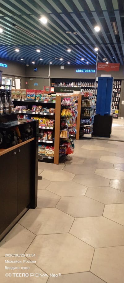 The media file is a photograph taken inside a store, likely a convenience store or a small supermarket, given the shelving stocked with various products. The scene is well-lit, suggesting it's daytime or the store has bright interior lighting. The floor is covered in large, beige hexagonal tiles, giving it a modern and clean appearance.

In the foreground on the left, there is a dark brown cabinet or counter with wooden accents. Above and behind it, shelves display bottles, possibly drinks or oils, and packaged snacks. Further back, more shelving units are visible, filled with a wide assortment of goods, including bags of chips or snacks, candy, and other food items. Some of the packaging has Russian text.

A prominent blue vertical structure, possibly a display or a piece of equipment, stands to the right of the product shelves. Above the shelves, signs with Russian text are visible on the wall and ceiling. One sign, in blue neon, reads "АВТОТОВАРЫ" (Autoparts), indicating a section of the store dedicated to automotive supplies. Another red sign says "ЭКСПРЕСС КАССА" (Express Checkout). There's also a sign with a green circle and Russian text that appears to be a store brand or section name, possibly "Азбука вкуса" (Alphabet of Taste).

The bottom of the image contains a timestamp and location information: "2026/06/26 03:54", "Можайск, Россия" (Mozhaysk, Russia), and "Введите текст" (Enter text), along with the device branding "TECNO POVA Neo 3". This information suggests the photo was taken in the early morning hours in Mozhaysk, Russia. The overall impression is of a retail environment with a variety of products for sale.

---
The media file is a photograph taken inside a retail store. The scene is brightly lit and shows rows of shelves stocked with various products. On the left, there are dark cabinets and shelves displaying bottles and packaged snacks. The main area features extensive shelving units filled with food items, candy, and other consumer goods. Prominent signs in Russian are visible, including "АВТОТОВАРЫ" (Autoparts) and "ЭКСПРЕСС КАССА" (Express Checkout). A blue vertical unit is also present in the aisle. The floor is covered in large, beige hexagonal tiles. The bottom of the image displays a timestamp "2026/06/26 03:54", the location "Можайск, Россия" (Mozhaysk, Russia), and a text prompt "Введите текст" (Enter text), along with camera branding. This suggests the photograph was taken in the early morning hours in Mozhaysk, Russia.