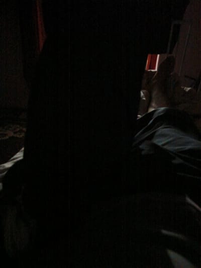 The image depicts a dimly lit interior scene, possibly a bedroom, at night. The central subject is a person who is largely obscured by shadow, with their face not clearly visible. They appear to be lying down, possibly in bed, with dark fabric draped over them. To the right of the frame, there is a glimpse of a person or an object that is also in shadow, with some indistinct shapes and textures visible. The overall impression is one of darkness and low light conditions, suggesting it is nighttime. There are hints of red or orange light in the background, possibly from a screen or a lamp, but these are not clearly defined. The setting appears to be indoors, with furniture and bedding hinted at by the textures and shapes. No text is visible in the image. The emotional tone is difficult to ascertain due to the lack of clarity, but the darkness and shadows create a sense of mystery or intimacy. The provided location context of Juma, Uzbekistan, offers a geographical setting for the image, but there are no specific visual cues in the image itself that directly confirm this location.