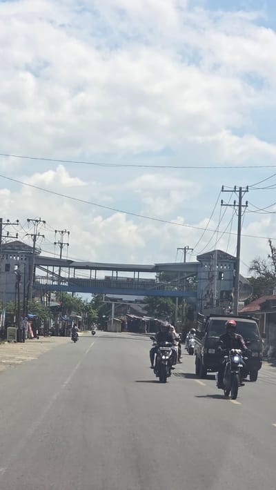 The image shows a street scene in Peub Lueng Nibong, Indonesia. A group of motorcyclists is riding on the right side of a paved road. A dark-colored vehicle is traveling with them. Overhead power lines and poles run along the side of the road. A pedestrian bridge spans the street. The sky is blue with scattered white clouds. Buildings and vegetation are visible on both sides of the road.