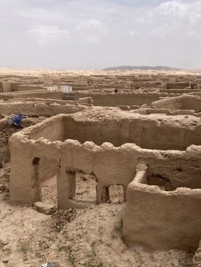 The image depicts an extensive view of the ruined city of Saranan, Pakistan. The scene shows numerous decaying mud-brick structures, mostly roofless, spread across an arid landscape. The sky is overcast, with a hazy horizon. In the background, elevated terrain can be seen. Some of the structures contain patches of water. A piece of blue fabric rests atop a wall towards the left-hand side of the image. In the middle distance, a figure in red is visible. The foreground is primarily sandy soil, with sparse vegetation.