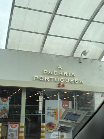 The image captures the exterior of "A Padaria Portuguesa" bakery, located in Palmela, Portugal. The establishment features a light-grey building facade with the name "A PADARIA PORTUGUESA" displayed in raised, light-colored letters. Below the name, the number "24" is visible in red, indicating 24-hour operation. Above the facade, a translucent, corrugated canopy supported by a metal framework extends outwards. A white, spherical light fixture or security camera is mounted on the upper right portion of the facade.

The ground level consists of large glass windows forming the storefront. Through these windows, several promotional posters are visible inside. One poster on the left displays bread items with a price of "2.45€". A central poster features a croissant and an orange beverage, accompanied by Portuguese text reading "VEIO ABASTECER? FIQUE PARA O PEQUENO-ALMOÇO." (Did you come to refuel? Stay for breakfast.) and a menu price of "4.45€". A double-door entrance is discernible, flanked by yellow and black striped bollards. The image is taken from inside a vehicle, with a portion of the vehicle's interior (dashboard/window frame) visible in the lower right, along with two rectangular papers affixed to the vehicle's windshield. The sky above the canopy appears overcast. No individuals are clearly identifiable within the frame.