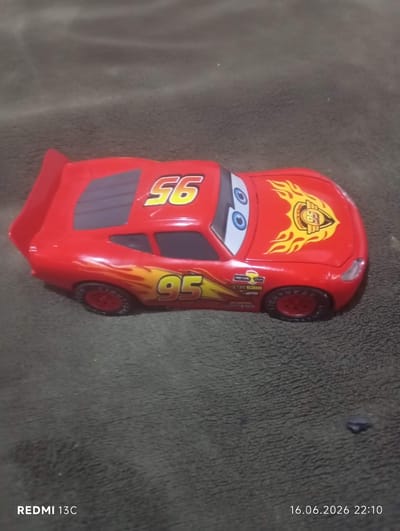 An overhead, slightly angled, close-up shot features a toy model of the animated character Lightning McQueen from the movie "Cars." The toy car is bright red with yellow flames along its sides and hood. The number "95" is prominently displayed on the side of the car, and the number "56" is on the roof. The car has large, cartoonish eyes on the windshield. The toy is positioned on a dark, textured surface that appears to be a blanket or fabric. The background is out of focus, with the lighting casting a soft glow on the toy. The lower left corner of the image displays the text "REDMI 13C", and the lower right corner shows the date and time "16.06.2026 22:10". The image does not contain any information about the city of At-Bashy, Kyrgyzstan.