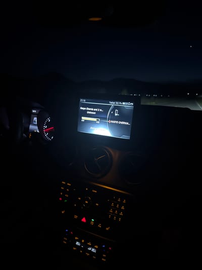 The image captures the illuminated interior of a car at night, likely in Voiniagovo, Bulgaria. The central focus is on the infotainment screen and the control panels below it.
The infotainment screen prominently displays "21:16", indicating the current time. It shows music playback information, including a title "Nasyo Chernia and 2 m..." by an "Unknown" artist, with track times "3:09" and "0:30". A phone call or contact named "NASYO CHERNIA..." is also visible. The screen indicates a "Yettel 5G Po" mobile network connection with full signal strength and a battery icon, along with "Dimitar's Phon" suggesting a Bluetooth-connected device.
Below the screen, two circular air vents are visible. Further down, the control panel features a phone keypad with numbers 1 through 9, 0, * and #, along with corresponding letters (ABC, DEF, GHI, JKL, MNO, PQRS, TUV, WXYZ). Various function buttons are present, including "RADIO", "MEDIA", "TEL", "NAVI", "CLEAR", "ON", "MODE", "AUTO", "ZONE", "OFF", "REST", and "A/C". Climate control displays show two temperature zones, one at "25°C" and another at "25" with an additional symbol. To the lower left, a partially visible red-illuminated instrument cluster, possibly a speedometer or tachometer, can be seen, along with a drive mode selector indicating "P" for Park. The interior lighting suggests the car is either parked or being driven in dark conditions.