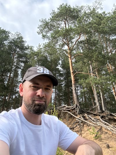 This is a selfie taken outdoors, oriented sideways. The subject is a man with a beard and a grey baseball cap with white lettering that reads "32". He is wearing a white t-shirt. The background consists of a pine forest with visible tree roots, and a sandy embankment. The sky is visible between the trees, with some clouds. The lighting suggests it is daytime, likely sunny. The setting appears to be a natural environment, possibly a park or forest area. The man's expression is neutral. No specific activity is depicted, other than the act of taking a selfie. The location context provided is Severnaya Samarka, Russia.