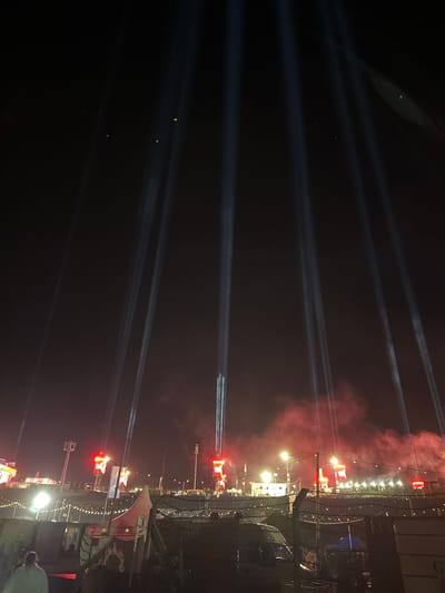 This image captures a nighttime scene at the Nürburgring in Nürburg, Germany, likely during a large event. The foreground shows a fence with some tents and vehicles behind it, suggesting a backstage or service area. In the mid-ground, there are structures with bright red lights and what appear to be large screens or illuminated signs. Beyond that, the background is dominated by a dark sky illuminated by numerous powerful white spotlights that shoot upwards, creating distinct beams of light. Red smoke or haze is visible in the air, particularly around the illuminated structures, indicating pyrotechnics or lighting effects. The overall impression is one of a concert or festival with elaborate stage lighting and a crowd atmosphere implied by the scale of the event.