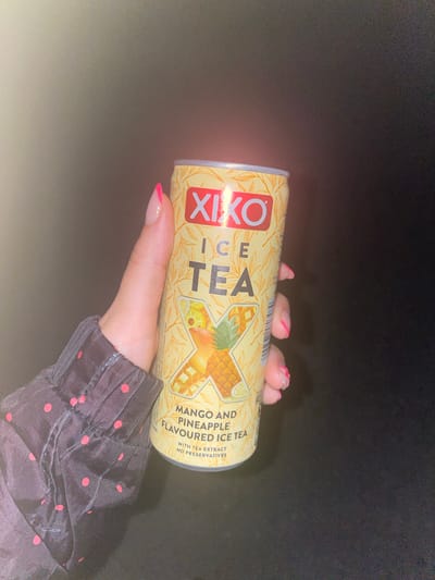 A close-up, dimly lit image taken in Kipilovo, Bulgaria, features a fair-skinned hand holding a 330ml Xixo Ice Tea can. The can is primarily yellow, with the brand name "XIXO" in red and "ICE TEA" in black. A large stylized "X" graphic on the can's label is composed of pineapple slices, mango cubes, and a lemon wedge. Text below the graphic indicates "MANGO AND PINEAPPLE FLAVOURED ICE TEA," with additional labels "WITH TEA EXTRACT" and "NO PRESERVATIVES." The hand displays a pink French manicure on the fingernails. A dark-colored sleeve with small red polka dots is visible on the left side of the frame, covering the wrist and forearm. The background is uniformly dark, with a subtle light source originating from the upper left, casting a slight glow.