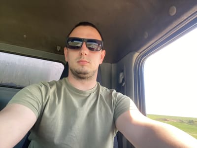 The image shows a man inside what appears to be a vehicle. He is wearing sunglasses and an olive-green shirt. The picture is taken from an unusual angle, making him appear upside down. Visible through the vehicle's windows is a landscape featuring a field. There is no evidence to link the image to Kapsal, Russia.
