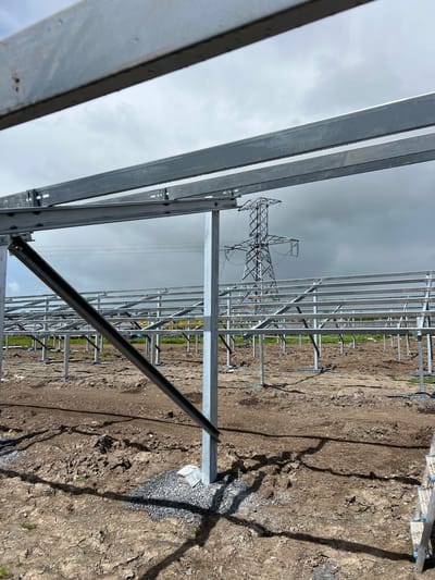 The image shows a partially constructed solar panel array in Grange Ville, Ireland. The skeletal metal framework of the array dominates the foreground and middle ground. In the background, an electrical transmission tower is visible against a cloudy sky. The ground is bare and appears recently disturbed, with some loose gravel present. There is no human presence evident in the image.