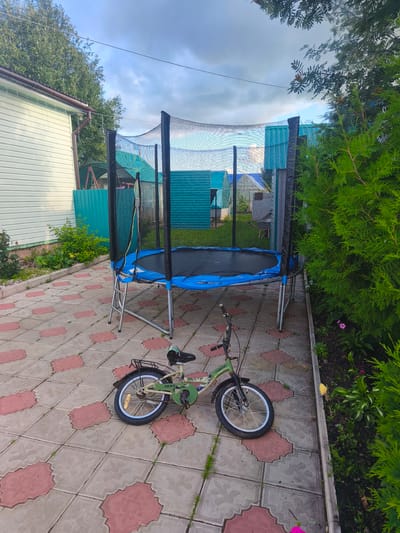 This is an outdoor, eye-level medium shot of a backyard scene in Burayevo, Russia. The primary subjects are a trampoline and a child's bicycle. The trampoline is large, round, and blue, with a black safety net enclosure. It is positioned on a paved patio. In front of the trampoline, a small green and beige bicycle with the word "ARMY" visible on its frame lies on its side. The patio is made of light grey interlocking pavers with red accents.

In the background, there is a light-colored house with a dark brown roof on the left. A turquoise fence runs along the property line. Further back, several small structures with turquoise roofs are visible, suggesting a rural or dacha-style setting. Lush green trees and bushes surround the yard, with a large evergreen shrub dominating the right side of the frame.

The scene appears to be set in the late afternoon or early evening. The sky is cloudy with patches of blue peeking through, indicating partly cloudy weather. There are no people visible in the image, and the overall mood is peaceful and still. The presence of the trampoline and bicycle suggests a place where children might play, but it is currently unused. The location cues, such as the style of the house and the surrounding vegetation, are consistent with a residential area in Russia.