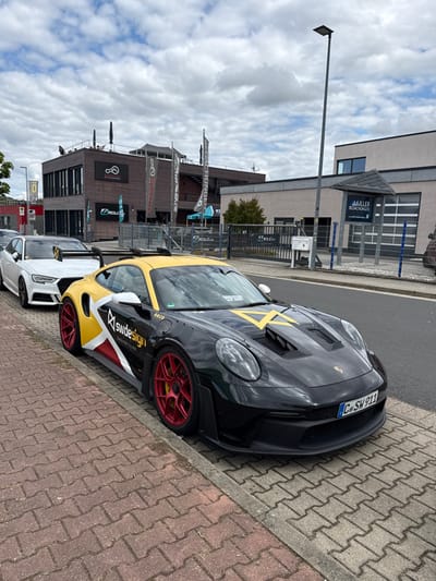The scene depicts a commercial street in Hohenstein-Ernstthal, Germany, under an overcast sky.
In the foreground and midground, a black Porsche 911, with a yellow roof, hood, and side accents, is parked on a reddish-brown brick paver sidewalk. The vehicle's side features "swdesign" branding in white text against a yellow, white, and red graphic. It is equipped with red multi-spoke wheels and yellow brake calipers. A German license plate displaying "C SW 911" is affixed to the front. Behind the Porsche, a white Audi sedan, also with a distinctive rear wing, is partially visible, parked on the same sidewalk. A grey concrete curb separates the sidewalk from an asphalt road.
The background features two primary buildings. To the left, a modern, multi-story structure with dark horizontal wood-paneled cladding displays a "ANALOG-WERK" logo and "Z MOLD" signage. This building has a rooftop terrace where several indistinct figures are seated under dark umbrellas. Multiple flags, some with "Z MOLD" and "DACHUNGEN" branding, are flown in front. To the right, a lighter-colored commercial building is visible, featuring a "MÜLLER BEDACHUNGEN" sign above a large garage door. A tall, grey street light pole stands in front of this building. A grey metal fence runs along the property line, separating the buildings from the street and sidewalk. Green foliage is visible between the buildings.
