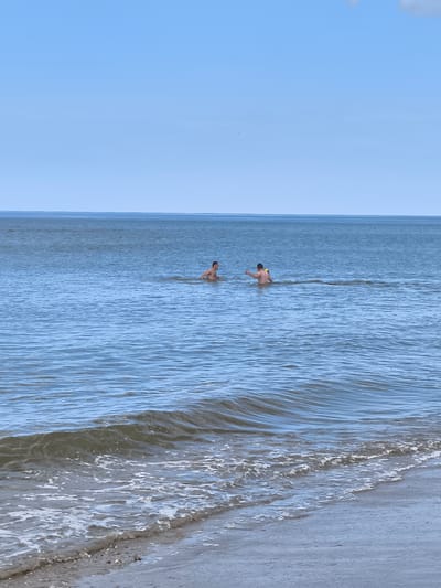The image shows two people swimming in the North Sea near Bergen, Netherlands. The foreground consists of the beach and gentle waves. The middle ground features the open sea with two individuals swimming. The background displays a clear, light blue sky. The weather appears calm and sunny.