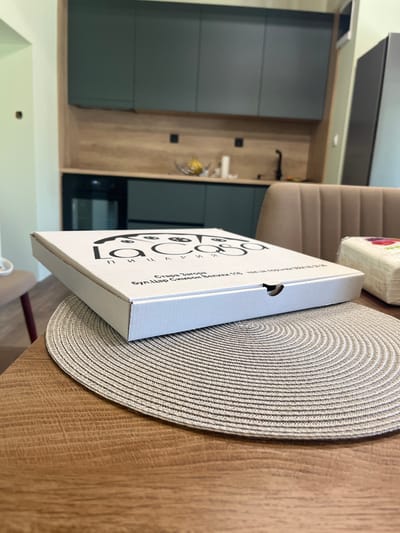 A white pizza box with a black logo that reads "la Cava" sits on a round, woven placemat on a wooden table. Text beneath the logo on the box reads "Стара Загора" (Stara Zagora), indicating the city in Bulgaria. Further text includes a street address and phone number in Bulgarian. In the background, a modern kitchen is visible with dark green cabinets, a wooden backsplash, and a black stovetop. To the right of the pizza box, a stack of white napkins with colorful floral patterns is visible. The scene appears to be indoors, likely in a home or restaurant setting.