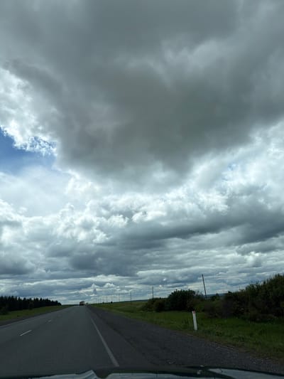 The image depicts a road in Горняк, Russia, under a sky filled with dramatic, layered clouds. The road is asphalt and appears to be a two-lane highway. The shoulder of the road is lined with green grass and vegetation. A few utility poles are visible along the roadside. The sky is dominated by various cloud formations, with some patches of blue sky peeking through.