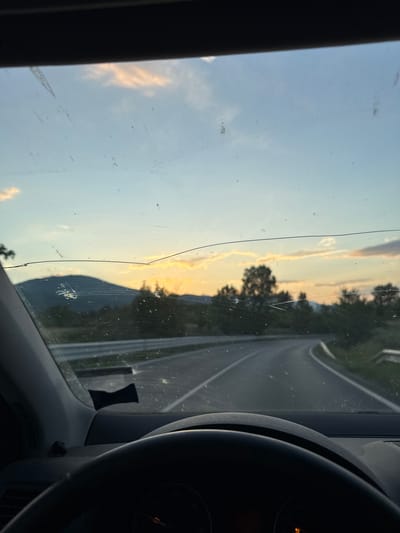 The image shows a view from inside a vehicle, possibly a car. A steering wheel is visible on the right side. The dashboard and front window frame are also visible. Through the windshield, a road is seen winding through a landscape with trees. The sky is visible, with some clouds and a hint of sunset colors. There is a crack on the windshield. The image provides no information regarding the location, subjects, objects, actions, interactions, or the environment of the city of Sadovo, Bulgaria.