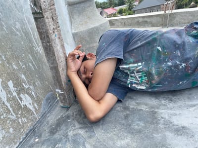 A male subject is observed lying on his left side on a concrete surface, identified as a rooftop, in Mns Asan, Indonesia. The subject is wearing a dark-colored t-shirt extensively splattered with various colors of paint. His eyes are closed, and his right hand is positioned near his face with fingers slightly curled, while his head rests on his bent left arm. A vertical wooden beam, exhibiting paint marks, is situated directly adjacent to the subject's head. The concrete rooftop surface displays cracks and scattered white paint splatters. In the background, beyond a low concrete barrier, a residential area is visible, featuring various houses with distinct rooflines and surrounding vegetation under natural light.