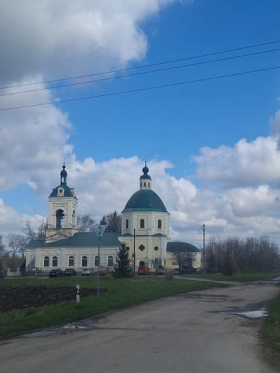 This image presents a serene daytime landscape, likely set in Паниковец, Russia, characterized by its architectural and natural elements. The main subjects are two prominent religious buildings, resembling Orthodox churches or cathedrals, with light-colored (possibly cream or light yellow) facades and distinctive green domes and spires. The structure on the right features a large central dome and a smaller spire, while the one on the left has a bell tower topped with a spire. Both buildings exhibit multiple arched windows.

In the foreground and midground, a dirt or gravel road runs along the right side of the frame, showing a visible puddle that suggests recent rain. Green grass lines the edge of the road and extends further into the scene. Bare deciduous trees, indicating late autumn, winter, or early spring, are scattered around the buildings and along the landscape. A few vehicles are faintly discernible parked behind the left-most building.

The sky is a bright blue, adorned with large, fluffy white and grey clouds. Several power lines stretch across the upper left section of the image. No people are visible, contributing to the quiet and undisturbed atmosphere of the scene. There is no visible text in the image.