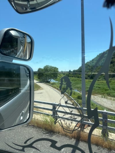 The image shows a view from a vehicle, likely a bus or truck, through the side mirrors and windshield. The scene depicts a road curving through a rural landscape under a clear blue sky. In the foreground, shadows of decorative metalwork are cast on the road and windshield. The side mirrors reflect the interior of the vehicle and the road ahead. A body of water, possibly a river, is visible alongside the road, with green vegetation and trees dotting the landscape. Hills covered in trees form the background. The location is identified as Ardhani, Greece.
