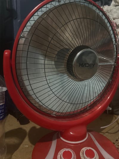 The image shows a close-up of a red electric fan, possibly a pedestal fan, with its grill and central motor housing prominently displayed. The fan is red with a white stripe on its casing. The motor housing has a silver-colored disc with some text on it, which appears to be "AIICVH". There are also two rotary switches on the fan's casing, one labeled with "OFF" and "HIGH", and another labeled with "OFF" and "OSCILLATE SWITCH". The fan appears to be indoors, and the background is somewhat blurred, suggesting the focus is on the fan itself. There are hints of a carpeted floor and what looks like some white fabric in the upper left corner. The lighting suggests an indoor setting, possibly during the day. No people are visible in the image. The setting could be a home or an office.