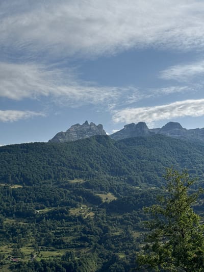 The image captures a panoramic view of a mountainous landscape with a clear blue sky dotted with scattered white clouds. In the foreground, a lush green forest covers the lower slopes of the mountains. Beyond the forest, rugged, rocky peaks rise sharply against the sky. The terrain appears to be in a rural or natural setting, with no visible structures or human activity in the immediate vicinity of the mountains. The overall impression is one of natural beauty and solitude. The location is identified as Redice, Montenegro.
