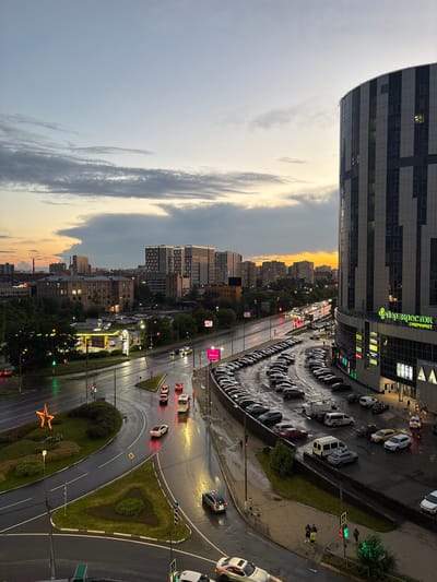 This is an aerial photograph of a street intersection and a parking lot at dusk in the city of Lesnye Polyany, Russia. The sky exhibits a gradient of colors from pale blue at the top to orange and gray clouds near the horizon, suggesting the sun has recently set. A large, modern, curved building with a dark facade and vertical white and gray panels dominates the right side of the frame. The building has a sign in Russian that reads "ПЕРЕКРЕСТОК СУПЕРМАРКЕТ".
The street is wet, indicating recent rain, and reflections of lights from buildings and vehicles are visible on the asphalt. Several cars are on the road, some appearing to be moving, while others are parked. Traffic lights are visible and illuminated, with one showing green.
A large, multi-row parking lot is situated in front of the curved building, filled with numerous cars of various colors and models, mostly parked. A smaller road with a star-shaped decorative light fixture on a green median is to the left, curving away from the main intersection. In the background, multiple apartment buildings and other structures are visible against the darkening sky, with some windows illuminated.
A few individuals are visible on the sidewalks and near the parking lot, appearing to be walking or standing. The overall scene depicts a commercial and residential urban environment during twilight, with active traffic and a busy parking area.