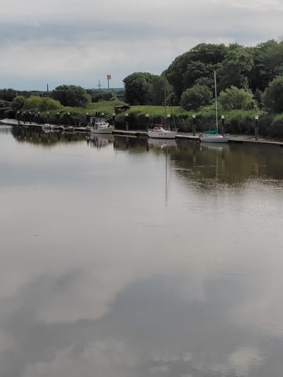 In Heiligenstedten, Germany, a calm river reflects the cloudy sky and a tree-lined bank. Several boats are moored along a wooden dock, including a white motorboat and two sailboats. One sailboat has a green sail hoisted, while another has a white hull and a red canopy. The background features lush green trees, a small building with a red roof, and a yellow sign on a pole. The water is still, with ripples creating a distorted reflection of the scene above. The overall impression is one of peacefulness and tranquility.