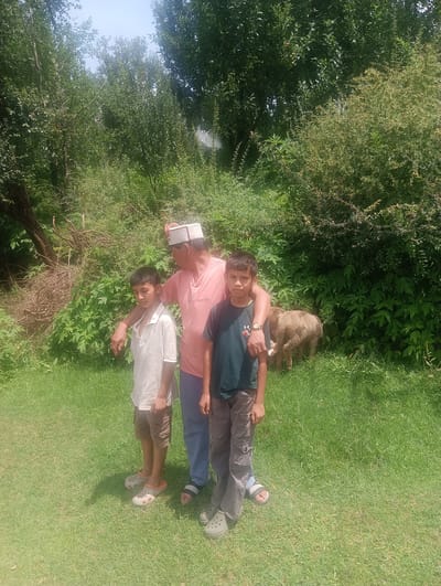The image depicts an outdoor scene with a man and two boys standing on a grassy area, with a sheep in the background. The man, wearing a pink t-shirt and a traditional cap, has his arm around the shoulder of the boy on the left. The boy on the left is wearing a white shirt and brown shorts. The boy on the right is wearing a dark t-shirt and grey pants. Behind them, there is dense green foliage and trees. A sheep is visible grazing in the grass. The location is identified as Khakhnal, India. The lighting suggests it is daytime.
