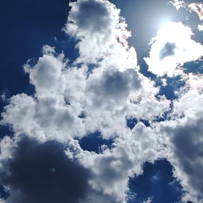The image is a photograph of a bright blue sky filled with various white and grey clouds. The sun is visible in the upper right quadrant, partially obscured by a large, bright white cloud. The clouds are varied in texture and density, with some appearing fluffy and wispy, while others are darker and more substantial, casting shadows. The overall impression is a dynamic sky with sunlight breaking through the clouds. There are no people, buildings, or geographical features identifiable in this image, nor is there any information that would allow for the determination of its specific location within the city of Торайғыров, Kazakhstan. The image is purely a skyscape.