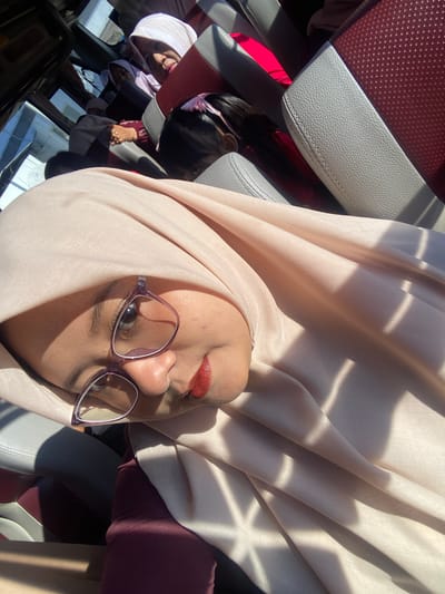 A young woman in a light peach hijab and purple-framed glasses is the primary subject of a selfie, taken inside a vehicle in Sumber Suko, Indonesia. She is looking towards the camera, with visible red lipstick. Her maroon-colored attire is partially visible beneath the hijab. Behind her, a woman in a white hijab smiles towards the camera. To her left, another individual wearing a light purple hijab is partially visible. The top of a child's head, adorned with a pink hair clip, is visible further back and to the right. The vehicle's interior features two-tone seats with light grey textured upholstery on the edges and perforated dark red material on the main sections. Strong direct sunlight streams into the vehicle, casting distinct geometric shadows across the main subject's hijab and the seat surfaces.