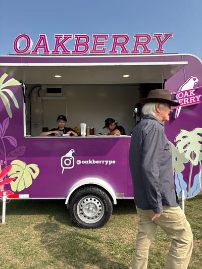The image depicts a bright, sunny day in Lurín, Peru, with a vibrant purple food truck named "OAKBERRY" as the central focus. The truck features large, stylized text spelling out its name in pink and white. A logo of a white bird on a pink circle, along with the Instagram handle "@oakberrytype", is visible on the side of the truck. Tropical leaf designs in shades of green and pink adorn the truck's exterior.
Two employees are visible inside the truck's service window. One, wearing a black baseball cap, is focused on something on the counter, possibly taking an order or preparing food. The other employee, also wearing a black cap, is partially obscured but appears to be working behind the counter.
In the foreground, an elderly man with white hair and a brown hat is walking past the food truck, his back mostly to the camera. He is wearing a dark, checkered long-sleeved shirt and light brown pants.
The food truck is parked on a grassy area, suggesting an outdoor event or location. The sky is clear and blue. The overall scene is casual and appears to be a typical day at a food vendor, possibly at a market or festival in Lurín.