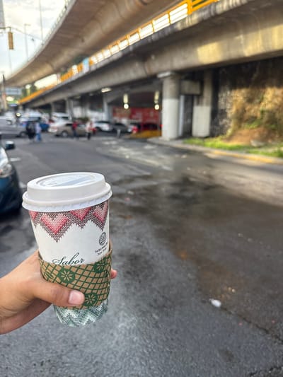 A person's hand holds a disposable coffee cup with a white lid and a decorative sleeve featuring a zig-zag pattern in red and pink, and a green patterned band with floral designs. The cup is held in front of a blurred urban background. In the background, a multi-level overpass structure is visible, with yellow lights lining its edge. Below the overpass, a street is wet and appears to have puddles. Several cars are parked or driving on the street, and some figures that appear to be people are discernible in the distance. The overall setting suggests an outdoor urban environment, possibly during daytime, with overcast or diffused lighting. The context provided indicates the location is in Tizapan, Mexico.
