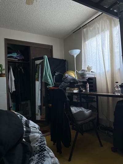 The image depicts a cluttered bedroom. A closet with sliding doors is visible on the left, containing clothes on hangers and shelves. A decorative cactus plant sits on top of a white cabinet next to the closet. In the center, a tall floor lamp stands beside a desk overflowing with various items, including books, containers, headphones, and a spray bottle. A chair is placed at the desk, with a light-colored cushion on it. A patterned blanket is spread out on a bed in the foreground, with a person partially visible. White sheer curtains cover a window on the right side of the room, where a desk is situated. The room appears to be in a residential setting, possibly an apartment or house. The image does not provide any information that would allow for the determination of its specific location within the city of Vancouver, United States of America.