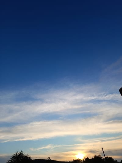 A serene landscape unfolds in Krasnopol'ye, Russia, under a vast expanse of deep blue sky, transitioning into lighter hues near the horizon. Wisps of delicate white clouds streak across the sky, creating a textured canopy. The lower portion of the image is dominated by a silhouetted tree line, indicative of rural or semi-rural surroundings. Among the trees, a lone utility pole stands, its wires stretching tautly across the scene. To the left, the sun, a brilliant orb of golden light, dips towards the horizon, casting a warm glow that illuminates the undersides of the clouds and hints at the fading light of day. The overall impression is one of tranquility and natural beauty.