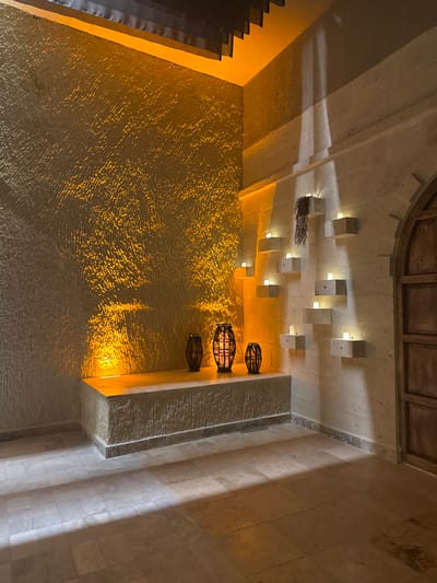 The image shows an interior architectural space with textured walls lit by warm, artificial lighting. The ceiling has a series of square, blocky light fixtures and three hanging, cage-like decorative elements. The space appears to be in Ürgüp, Turkey, given the style of architecture and lighting. No people are visible.
