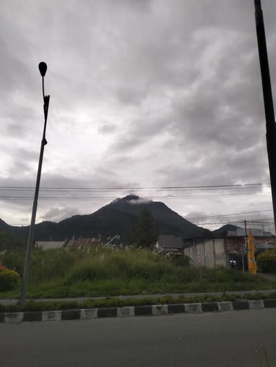 The image depicts a scene in Panteraya, Indonesia, under an overcast sky. In the foreground, a paved road runs horizontally across the bottom of the frame, bordered by a curb marked with alternating black and white blocks. To the left of the road, a tall lamppost stands silhouetted against the sky. Beyond the curb, a grassy area is visible, with tall, dry reeds or grasses interspersed with greener vegetation.

In the middle ground, several buildings are present. One building has a weathered, light-colored facade, and another appears to have a reddish roof. Signs are visible on some of the structures; one prominent yellow banner with Indonesian text is affixed to a building on the right. A circular sign with the number "30" inside a red border is also visible on a wall. Power lines are strung across the scene, supported by poles.

In the background, a large, dark mountain range dominates the horizon, with its peak shrouded in clouds or mist. The overall atmosphere is muted, consistent with cloudy weather. No people or animals are clearly visible in the image. The scene appears to be along a roadside in a semi-urban or suburban area, adjacent to a natural landscape.