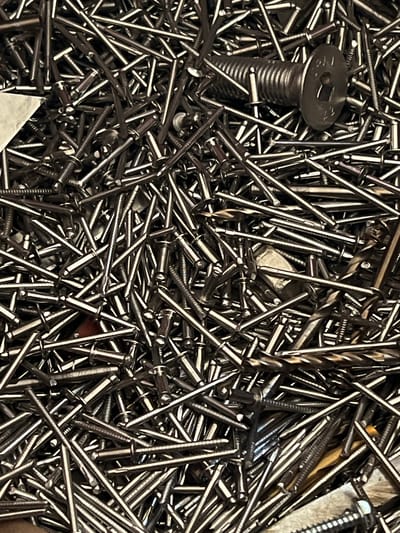 The image displays a large pile of assorted metal fasteners, predominantly nails and rivets. A single larger bolt with a hexagonal recess in its head is prominently visible near the top center. The fasteners are metallic, appearing to be made of steel or a similar alloy, and are scattered randomly, creating a densely packed composition. The lighting suggests an indoor setting, possibly a workshop or storage area. No people or specific environmental features that would indicate a location in Lanžhot, Czechia are discernible within the image.