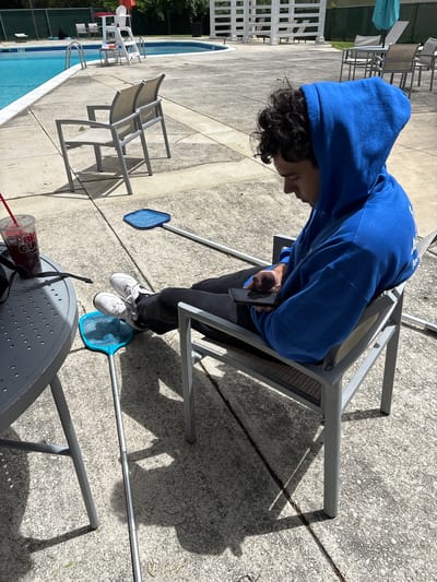 A person in a blue hoodie is sitting on a chair near a swimming pool. They are looking at a cell phone in their hands. Two pool skimmers are on the ground near the chair. A table with a drink on it is also nearby. The location appears to be an outdoor pool area, possibly a residential or community pool. The city is mentioned but not visible in the image.