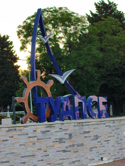The image shows a large, blue, three-dimensional sign that reads "TUAPSE" in Russian Cyrillic letters. The sign is decorated with elements suggestive of a seaside or port town, including a ship's wheel and two stylized white birds, likely representing seagulls, in flight. The letters themselves have a textured, patterned design in shades of purple and pink.
The sign is mounted on a low, multi-layered stone wall, constructed from rectangular blocks of varying shades of gray, beige, and tan. Behind the sign, lush green trees form a dense backdrop, with the tops of several tall evergreen trees visible on the left and right edges of the frame. A bright sky, appearing overcast or at dusk, is visible in the upper left portion of the image. Sunlight can be seen filtering through the trees on the left, casting a warm glow.
In the background, beyond the sign and the stone wall, a white ornate fence can be seen, suggesting a park or public space. A lamppost is also visible behind the sign. The overall scene evokes a welcoming atmosphere, characteristic of a tourist destination. The time of day appears to be late afternoon or early evening, based on the soft lighting and the hint of sunset colors through the trees. The weather seems calm and pleasant, with no indication of harsh conditions. The presence of the ship's wheel and seagulls strongly suggests a coastal location, consistent with Tuapse being a port city on the Black Sea. No people are visible in the frame.