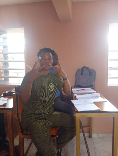 The scene takes place in an interior room in Benin City, Nigeria, appearing to be a classroom or study area. A dark-skinned individual with short, twisted hair, dressed in a green patterned two-piece ensemble featuring a circular logo on the chest and thigh, is seated in a wooden chair with a gold-colored frame, facing the camera. This individual is performing a bilateral hand gesture, extending the index, middle, and pinky fingers while raising the thumb on each hand. Their mouth is slightly open. Behind this person, another dark-skinned individual wearing a blue shirt is partially visible, obscured by the foreground figure. To the right of the foreground person, a wooden desk with a gold-colored metal frame holds a grey backpack, multiple white papers, and a red pen. A partial view of another wooden desk with papers is visible on the left. The room is characterized by light peach-colored walls and two windows with horizontal blinds, through which bright natural light enters. A ceiling beam is present.