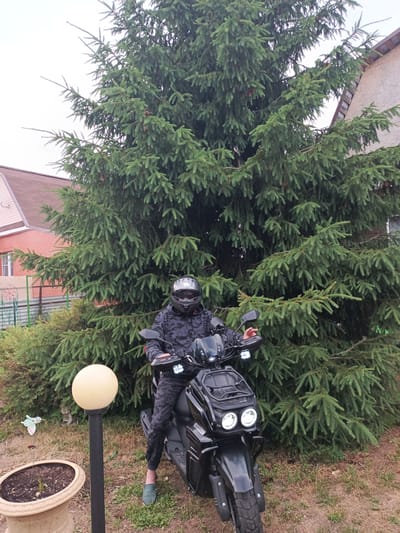 A person wearing a camouflage jacket, camouflage pants, and a black helmet with a clear visor sits on a black scooter in front of a large evergreen tree. The scooter has two headlights and a front rack. To the left of the scooter, a round, yellow outdoor light stands on a black pole, next to a tan planter with soil and a small plant. In the background, there is a red brick house with a brown roof and a section of a lighter-colored building attached to it. A metal fence is visible behind the house. The ground is covered in grass and dry leaves, and the sky is overcast. The location is identified as Novoye, Russia.